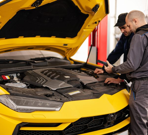 Two mechanics stand with tbalet against open hood of yellow sport car suv.